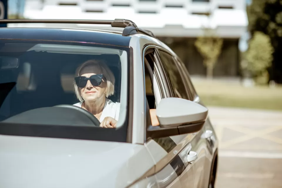 Senior woman wearing sunglasses sitting in the driver's seat of a silver car.
